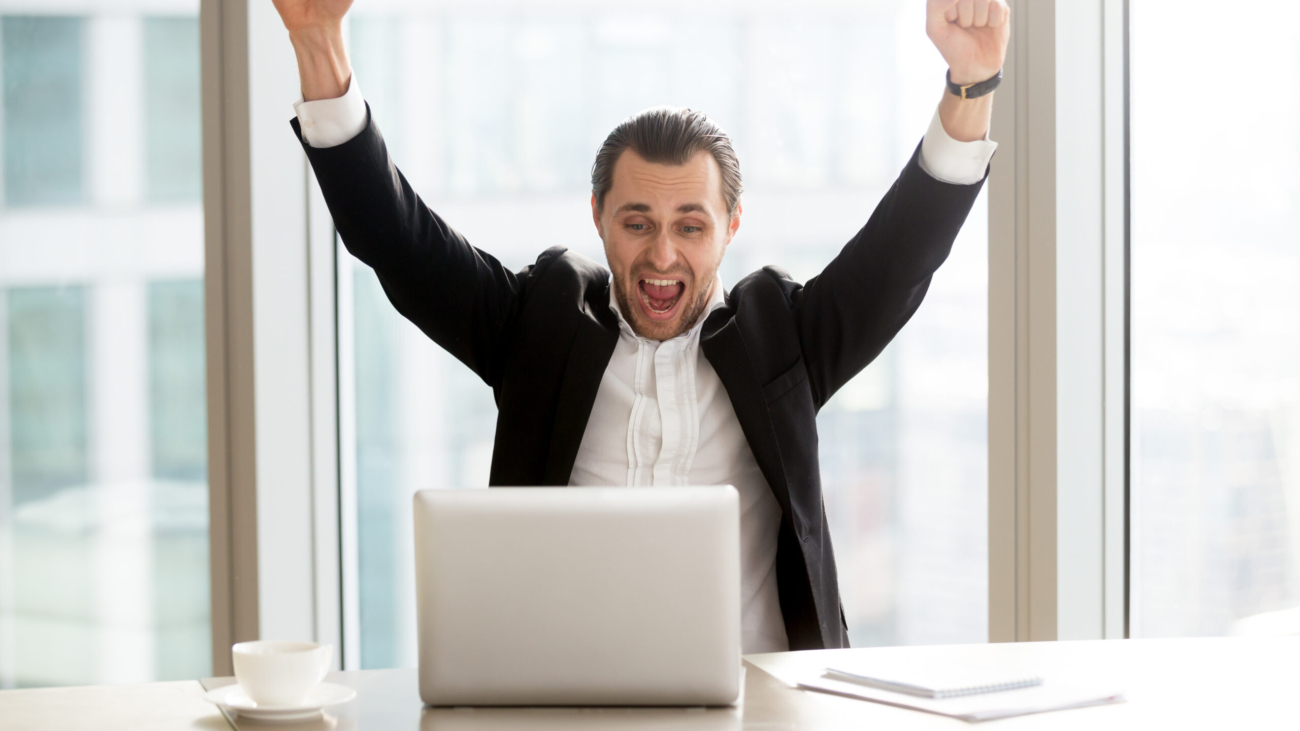 Happy handsome businessman in front of laptop at desk in modern office setting celebrating great news, screaming in joy because of victorious achievement. Successful deal, business success concept.